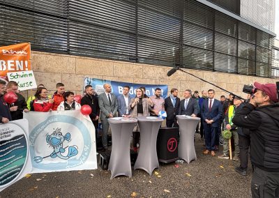 Eine Frau spricht in ein Mikrofon vor dem Banner „Bäder in Not“, umgeben von weiteren Teilnehmenden und einem Kamerateam.