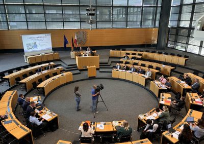 Blick in den Plenarsaal mit Teilnehmenden, Kamerateam und Präsentation „Thüringer Bäder in Not“.